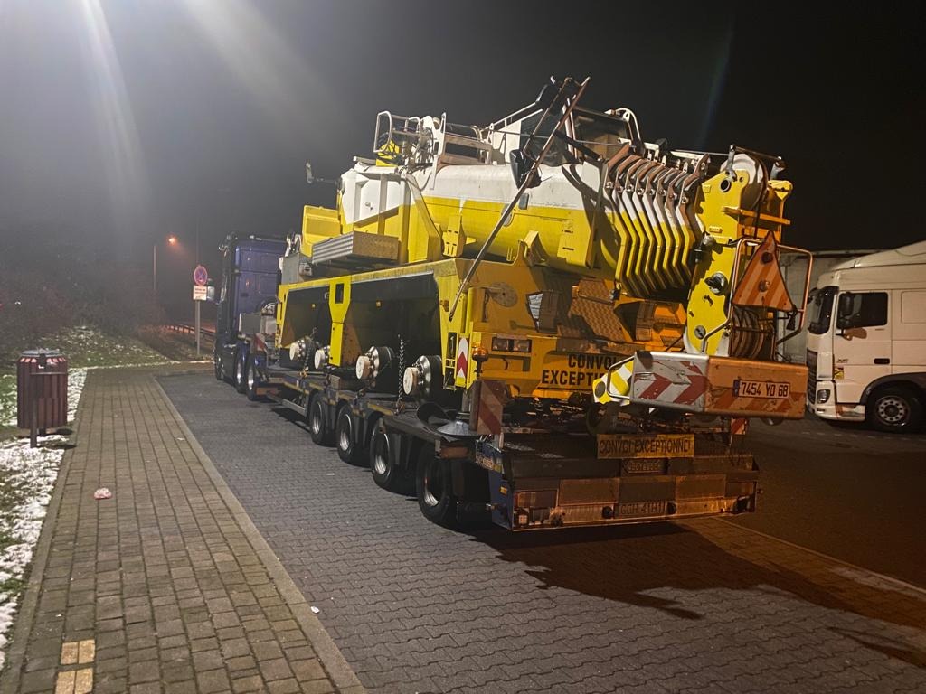 Nocny transport ponadgabarytowy: Żółty dźwig na niskopodwoziowej platformie z tablicą 'Convoi Exceptionnel' i francuską rejestracją, zaparkowany na oświetlonym parkingu dla ciężarówek.
