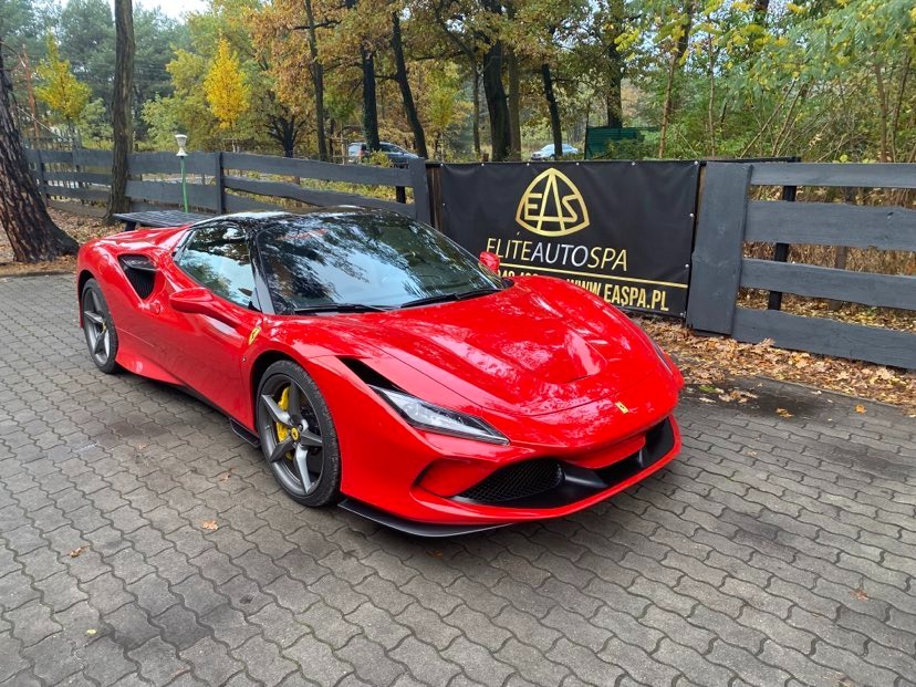 Czerwone Ferrari F8 Spider zaparkowane na kostce brukowej przed ogrodzeniem i banerem Elite Auto Spa, widoczne żółte zaciski hamulcowe.