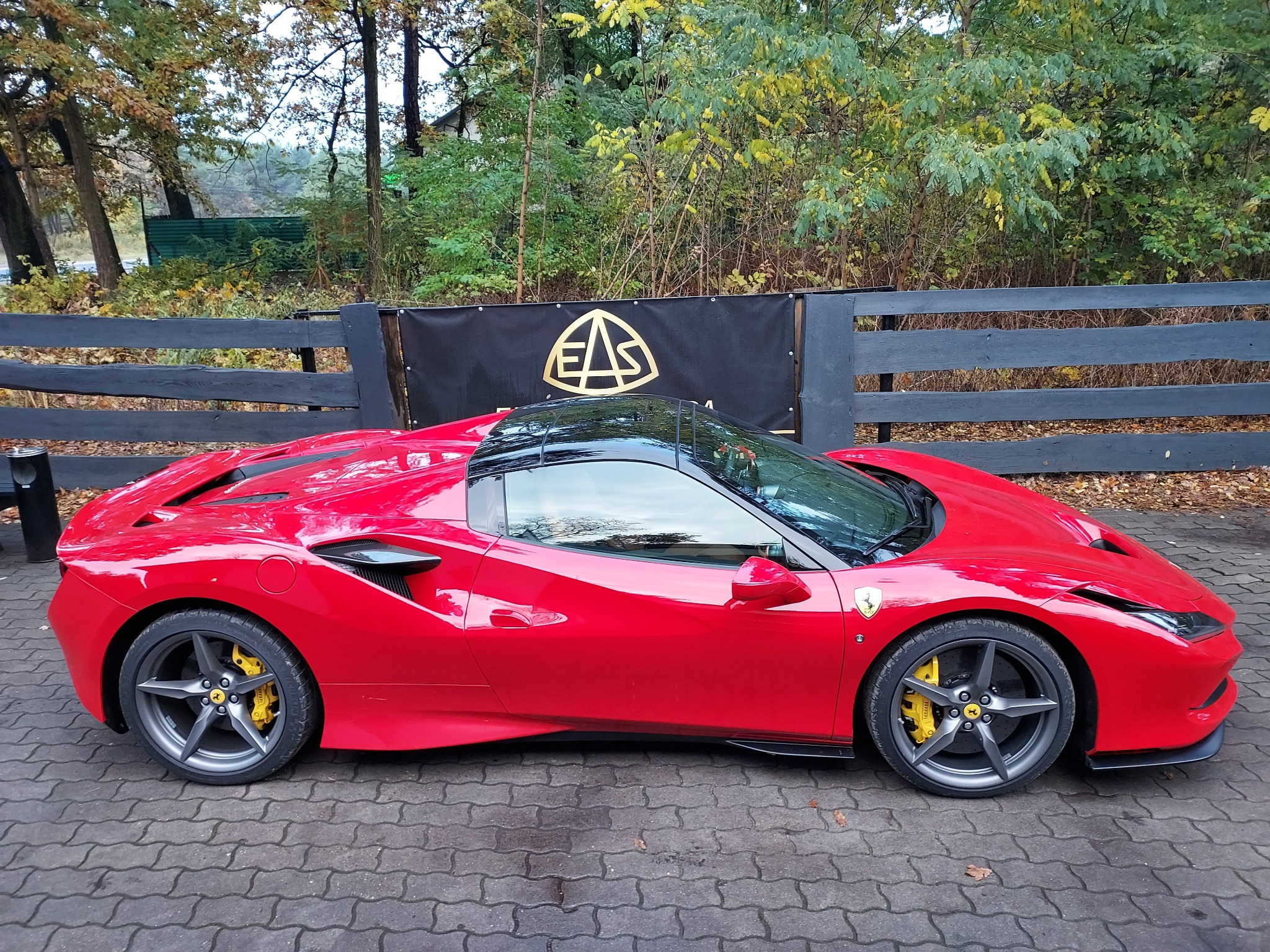 Czerwone Ferrari zaparkowane na brukowanej powierzchni, z żółtymi zaciskami hamulcowymi i szarymi felgami, na tle czarnego płotu z logo EAS i zieleni drzew.