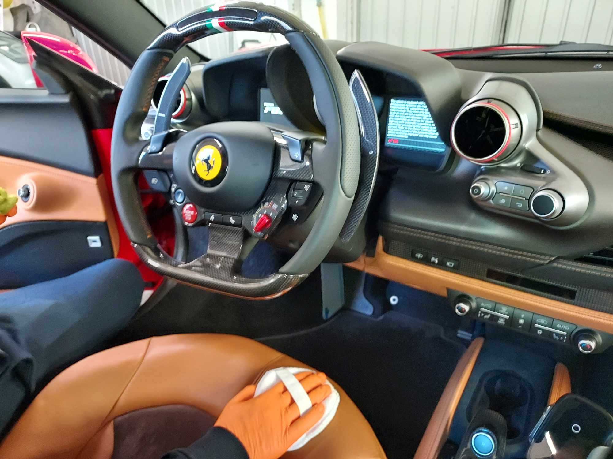 Wnętrze czerwonego samochodu Ferrari z brązową skórzaną tapicerką, czyszczone przez osobę w pomarańczowej rękawicy za pomocą białej ściereczki.