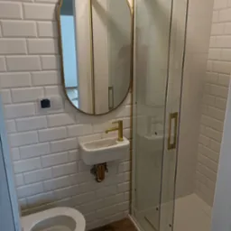 Nowoczesna łazienka z białymi, prostokątnymi płytkami, owalnym lustrem w złotej ramie, małą umywalką z baterią w kolorze złota i kabiną prysznicową ze złotymi detalami, widoczny fragment toalety.