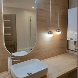 Łazienki & Remonty Cabanek - Nowoczesna łazienka z umywalką nablatową, owalnym lustrem z podświetleniem LED, drewnopodobną okładziną ścienną i wiszącymi lampami kulami, z widocznym piecem gazowym w rogu.