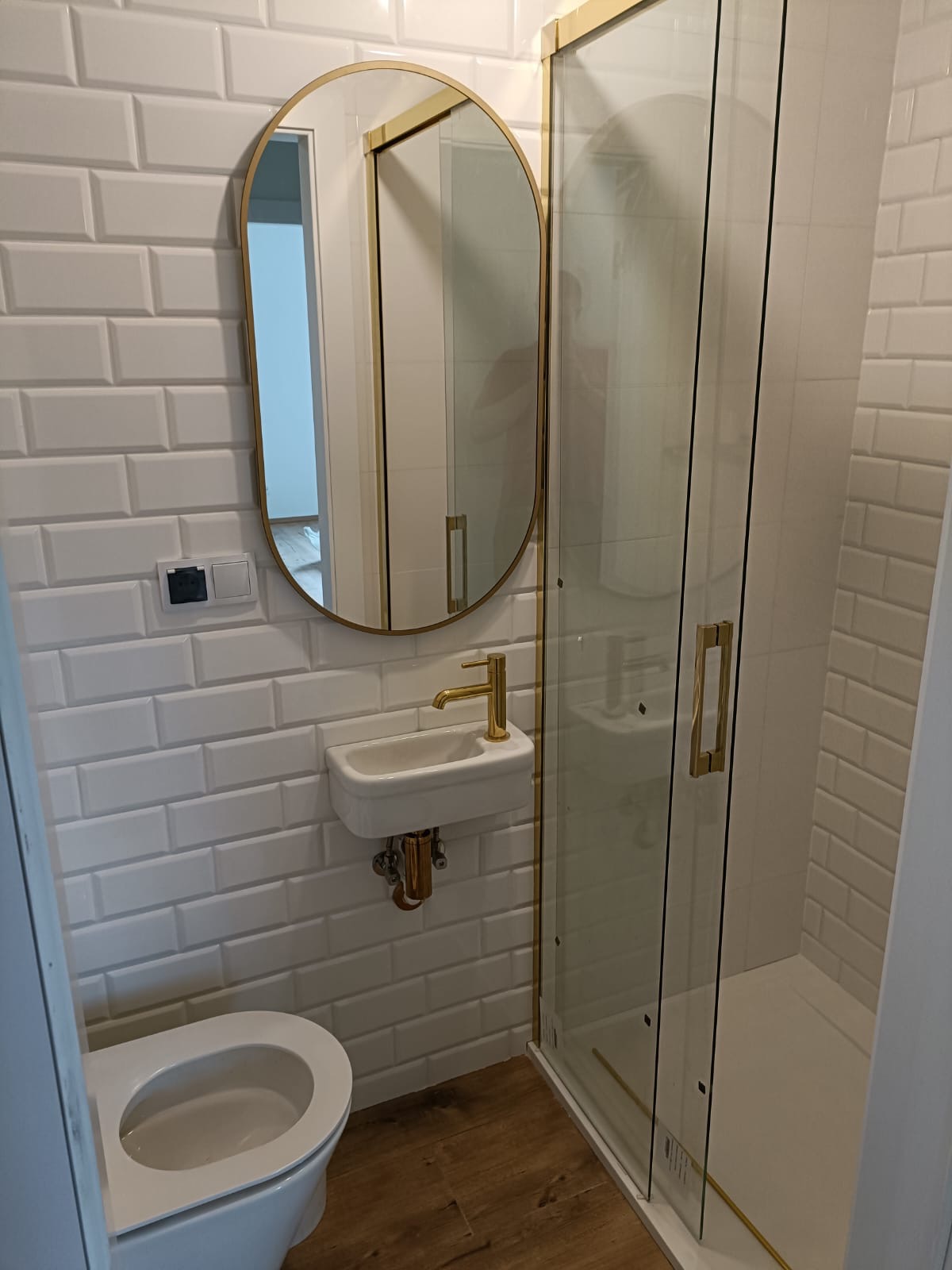 Nowoczesna łazienka z białymi, prostokątnymi płytkami, owalnym lustrem w złotej ramie, małą umywalką z baterią w kolorze złota i kabiną prysznicową ze złotymi detalami, widoczny fragment toalety.