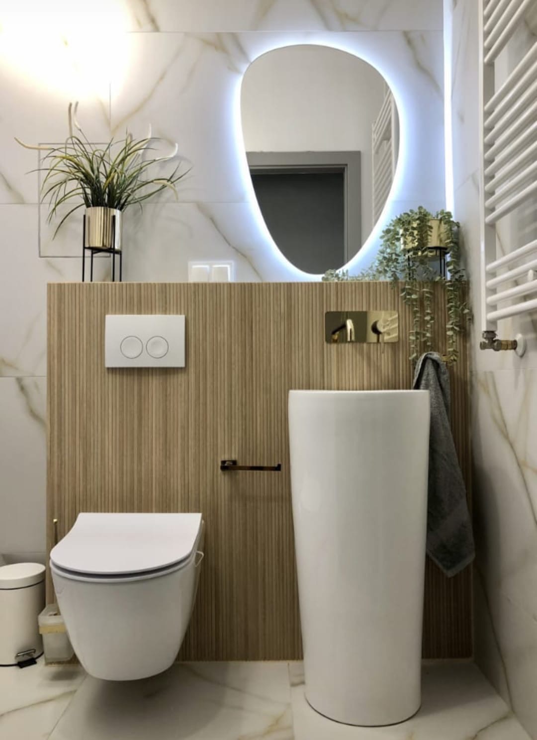 Nowoczesna łazienka z marmurowymi płytkami, podświetlanym lustrem o nieregularnym kształcie, wiszącą toaletą i wolnostojącą umywalką, uzupełniona o złote akcenty i rośliny doniczkowe.