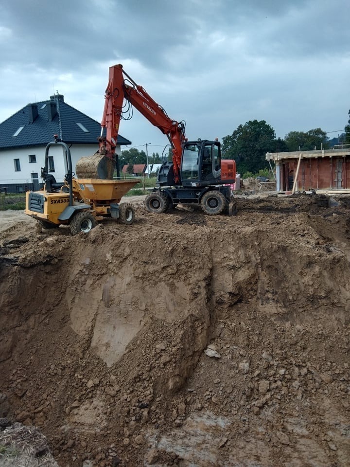 Żółta wywrotka SXR500 i pomarańczowa koparka Hitachi na placu budowy, świeżo wykopana ziemia, w tle dom z ciemnym dachem i budynek w trakcie budowy z czerwonej cegły.