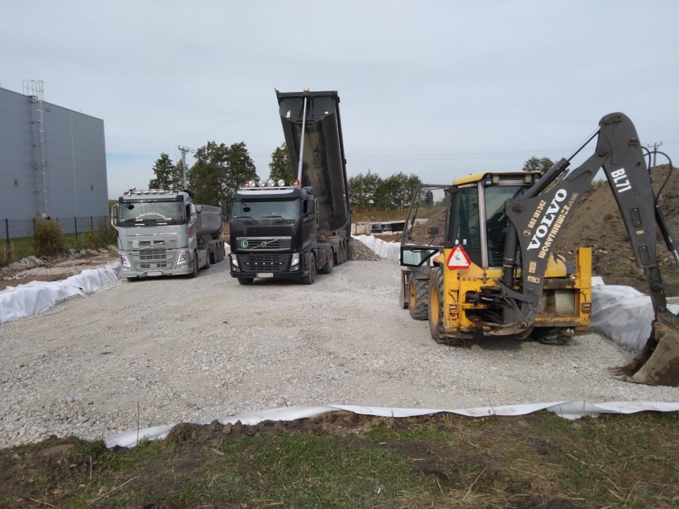 Dwa samochody ciężarowe Volvo i żółta koparko-ładowarka Volvo BL71 na placu budowy z utwardzoną, żwirową powierzchnią i białym materiałem geowłókninowym na obrzeżach.