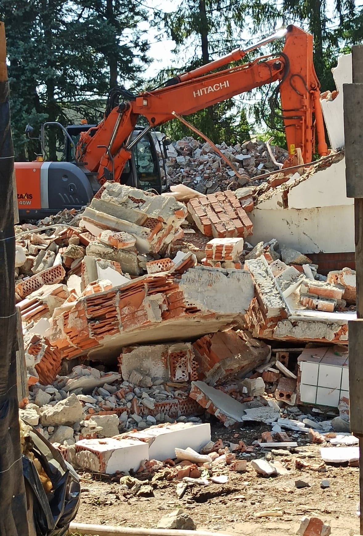 Pomarańczowa koparka Hitachi Zaxis 40W na tle gruzowiska z cegieł i betonu po rozbiórce budynku, widok zza fragmentu ściany.