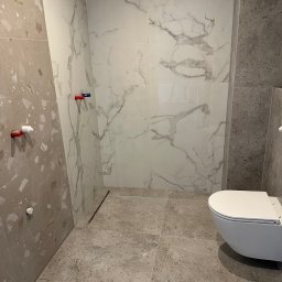 Elite painting - Nowoczesna łazienka z marmurowymi płytkami ściennymi i szarą terakotą na podłodze, widoczny odpływ liniowy i wisząca toaleta, surowe przyłącza wodne na ścianie.