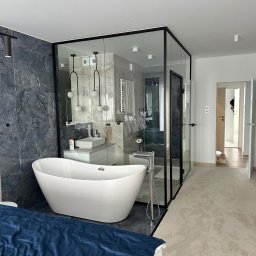 Elite painting - Nowoczesna łazienka z wolnostojącą białą wanną, kabiną prysznicową ze szklanymi ścianami w czarnej ramie, umywalką na szafce i szarą ścianą z marmurowym wzorem, widok z sypialni na wykładzinę...