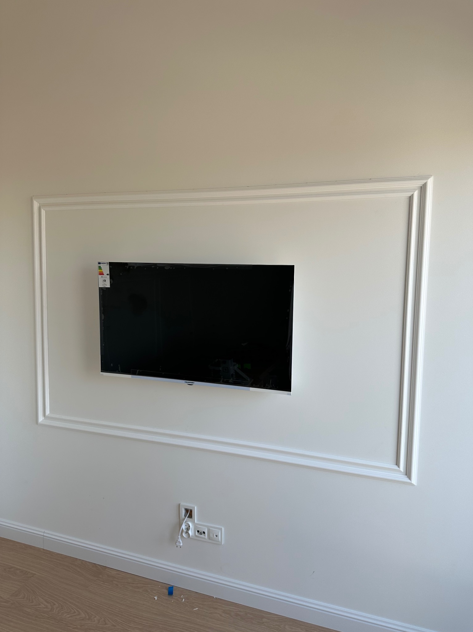 Telewizor zamontowany na ścianie w ozdobnej ramie z listew dekoracyjnych, widoczne gniazdka elektryczne i fragment podłogi z paneli.