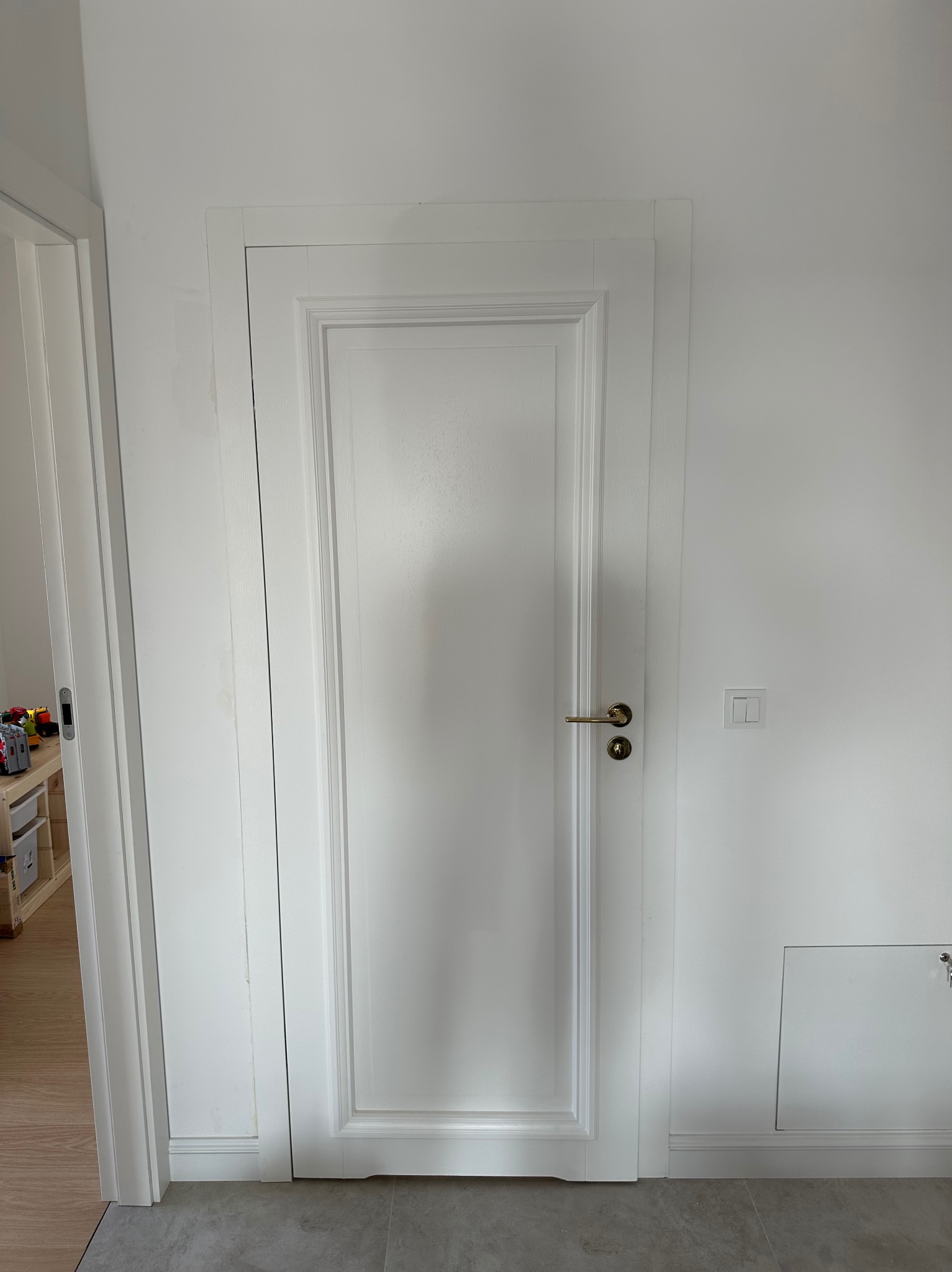 Białe drzwi wewnętrzne z ozdobną ramą i złotą klamką w nowo wykończonym wnętrzu, widoczny włącznik światła i kluczyk do szafki rewizyjnej.