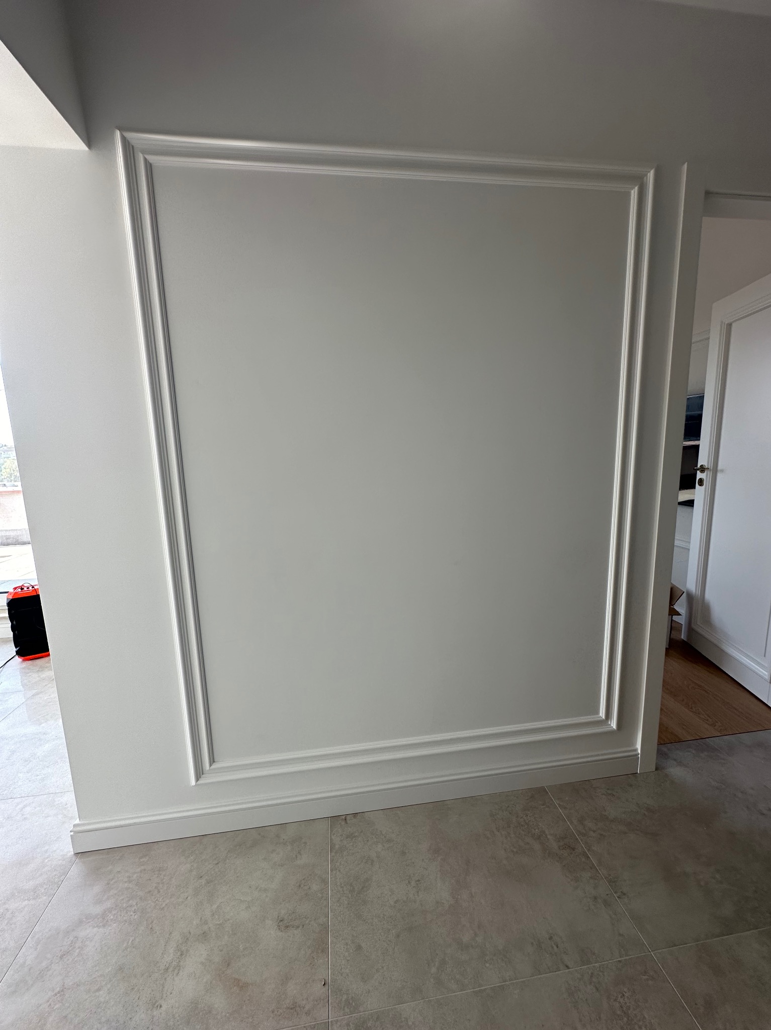 Ściana z dekoracyjną, białą sztukaterią w kształcie prostokąta, dolna listwa przypodłogowa, płytki podłogowe imitujące kamień, widoczny fragment grzejnika elektrycznego w tle.