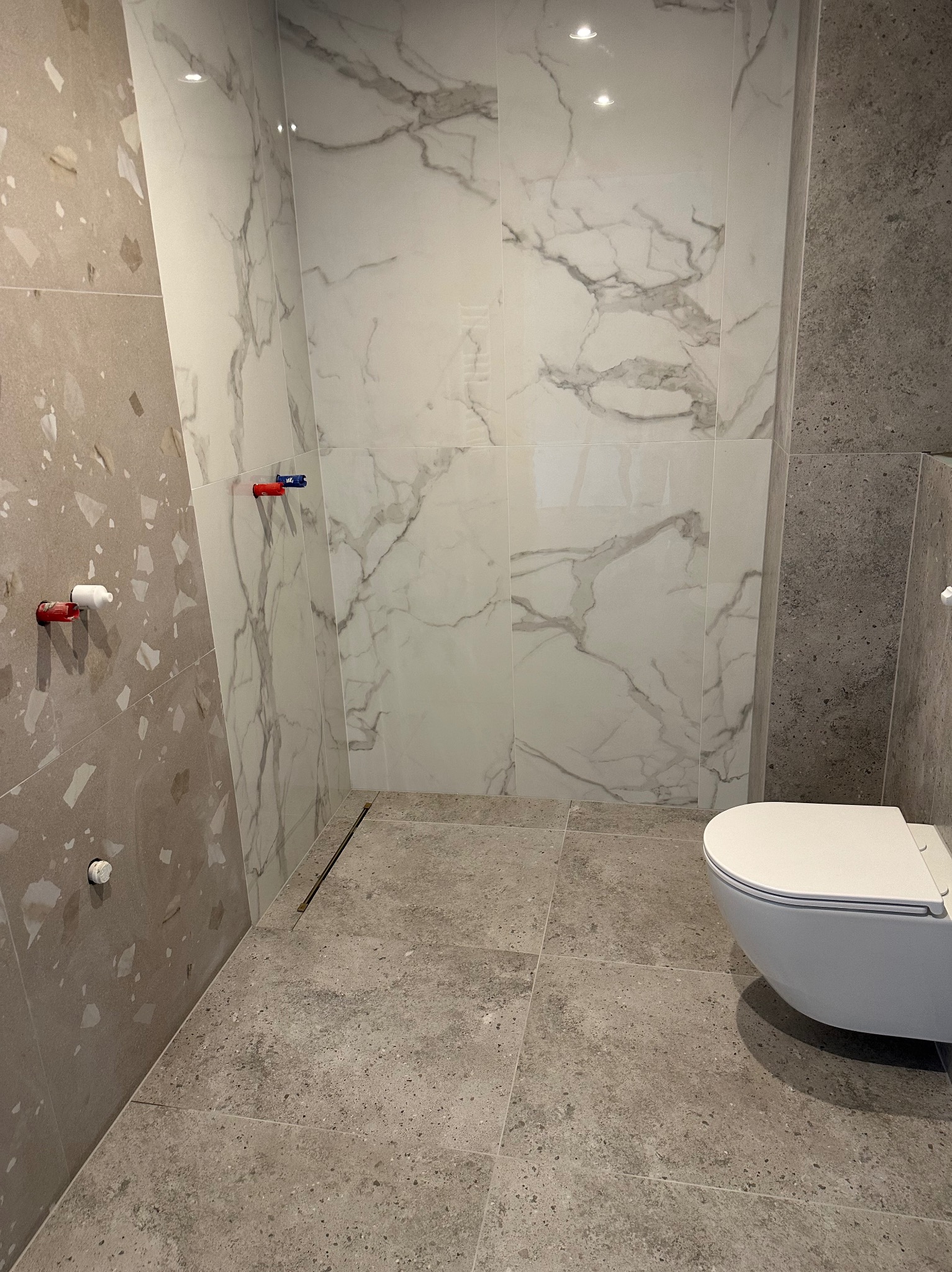 Nowoczesna łazienka z marmurowymi płytkami ściennymi i szarą terakotą na podłodze, widoczny odpływ liniowy i wisząca toaleta, surowe przyłącza wodne na ścianie.