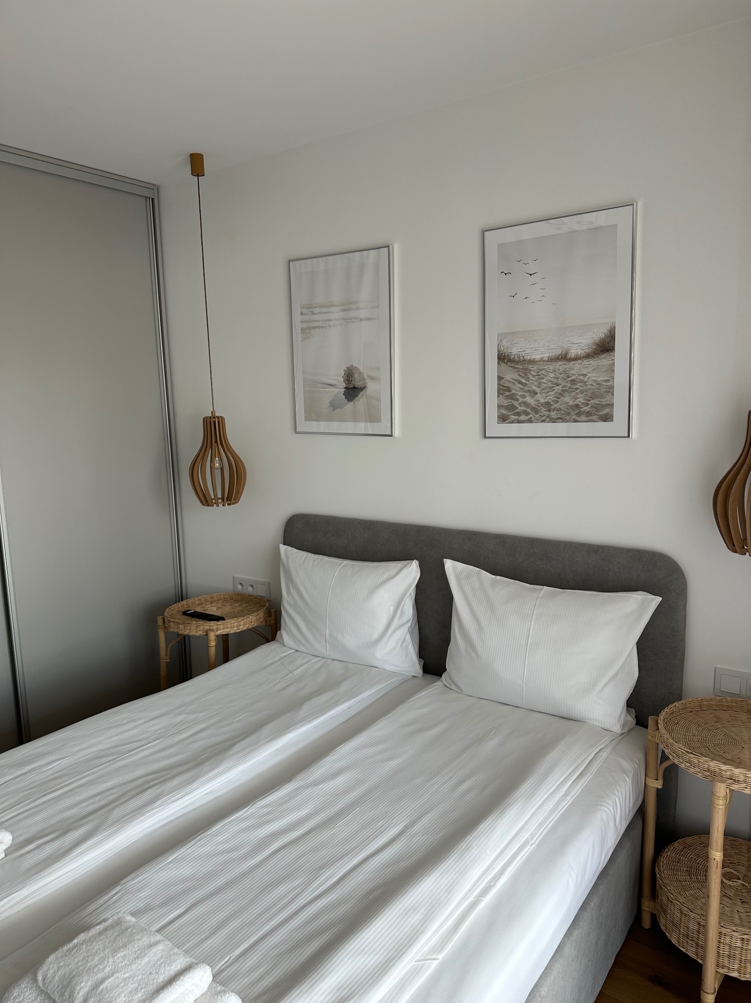 Jasna sypialnia z dwoma łóżkami, szarą ramą i białymi pościelami, ozdobiona drewnianymi lampami wiszącymi i obrazami z motywami plażowymi, widok na szafę przesuwną.