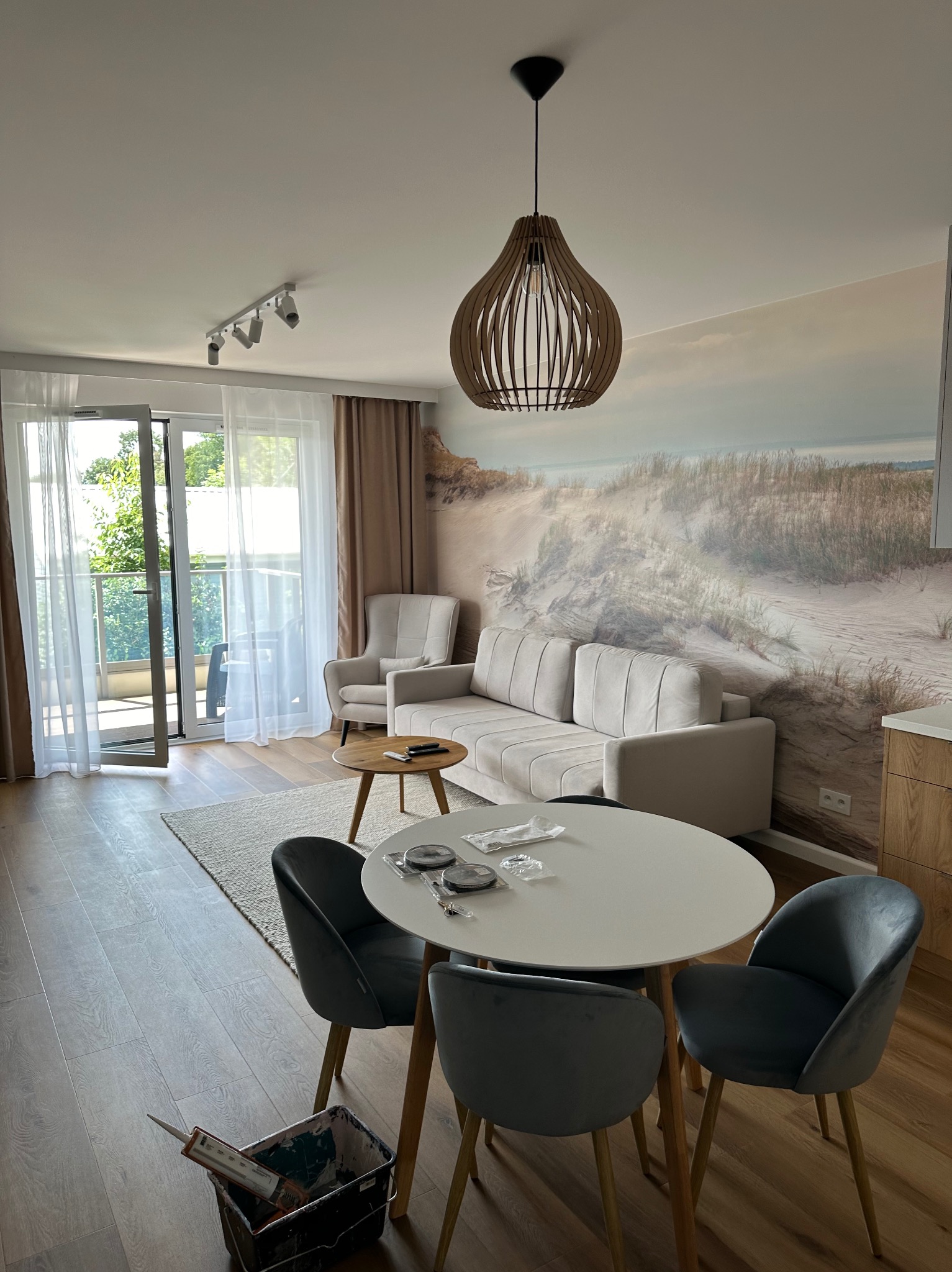 Salon po odświeżeniu: fototapeta z motywem wydm, drewniana lampa wisząca, szare krzesła welurowe przy okrągłym stole, otwarte drzwi balkonowe z widokiem na zieleń, wiadro z narzędziami malarskimi...