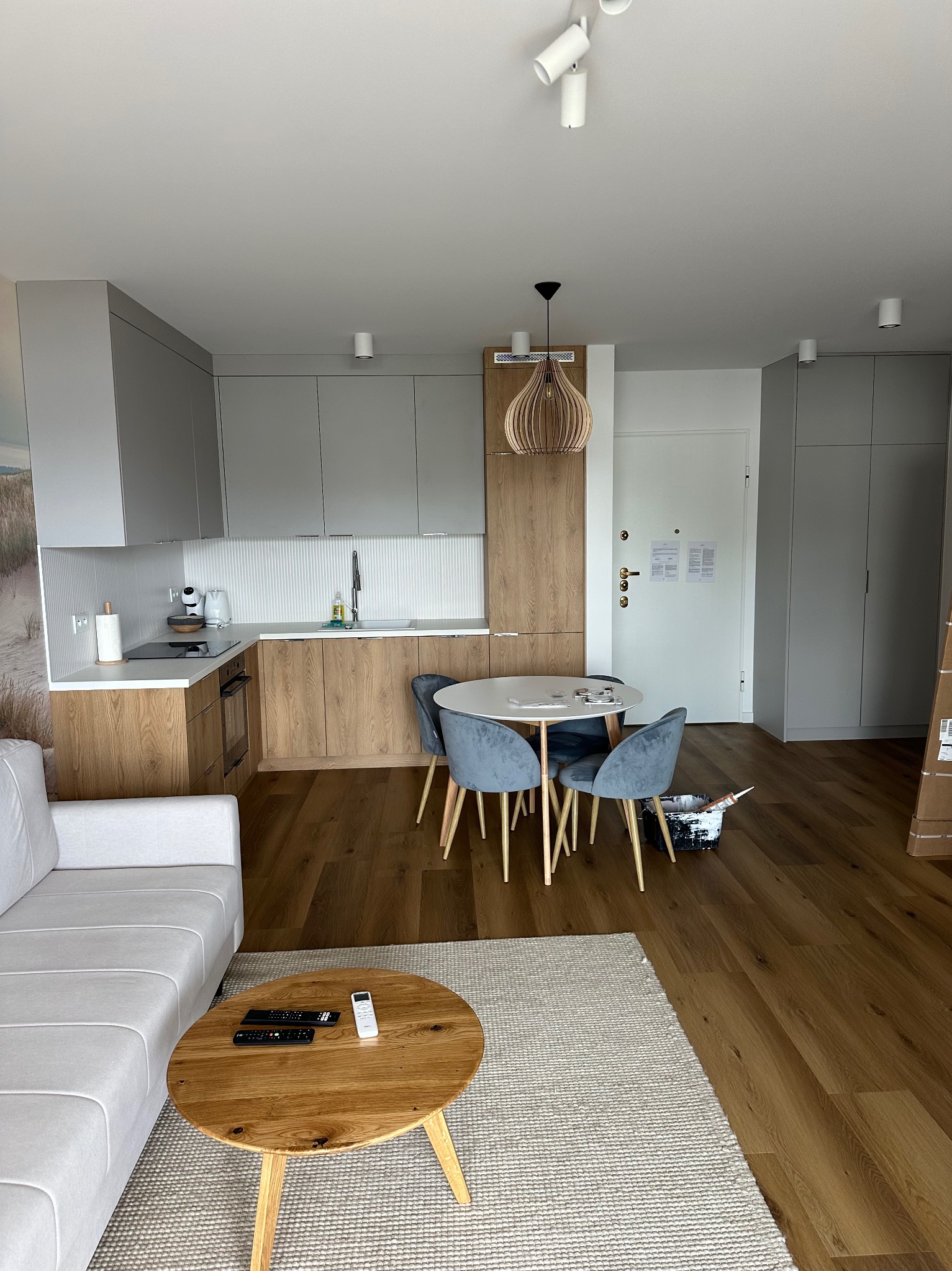 Jasne wnętrze mieszkania: drewniana podłoga, szara zabudowa kuchenna z drewnianymi akcentami, okrągły stół z szarymi krzesłami, biała sofa i okrągły stolik kawowy z pilotami. Widoczny kubeł z farbą...