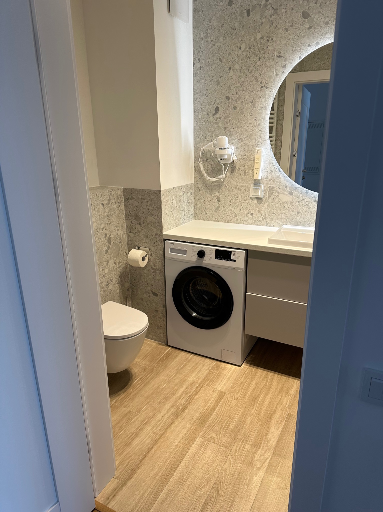 Nowoczesna łazienka w odcieniach szarości i beżu z okrągłym podświetlanym lustrem, pralką pod blatem i wiszącą toaletą.