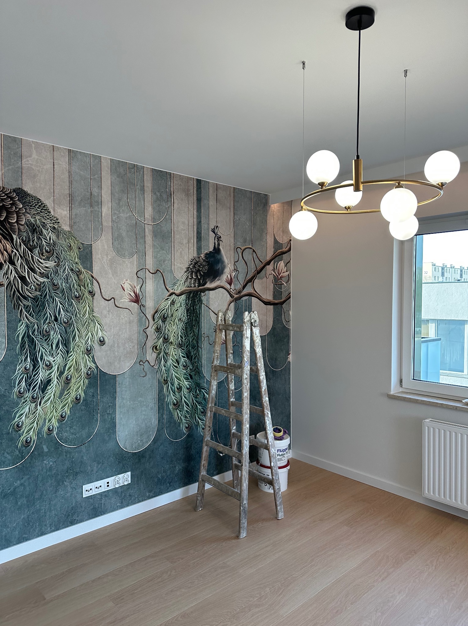 Ściana z fototapetą przedstawiającą pawie, drabina malarska i puszki farb w pokoju z parkietem i nowoczesną lampą.