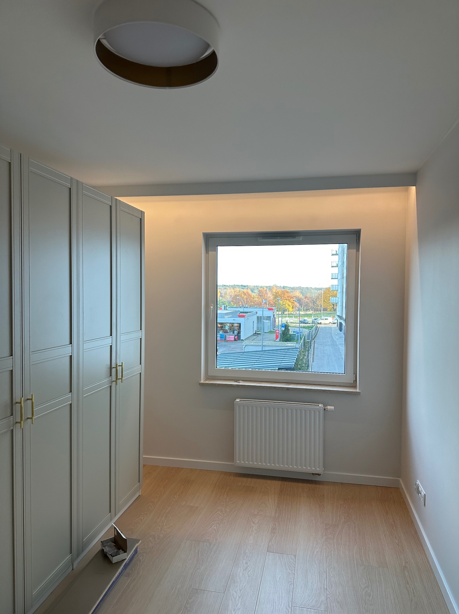 Wykończone wnętrze pokoju z szafą w zabudowie w kolorze szarym, widokiem na zewnątrz przez okno i panele na podłodze. W tle widoczny budynek i drzewa.