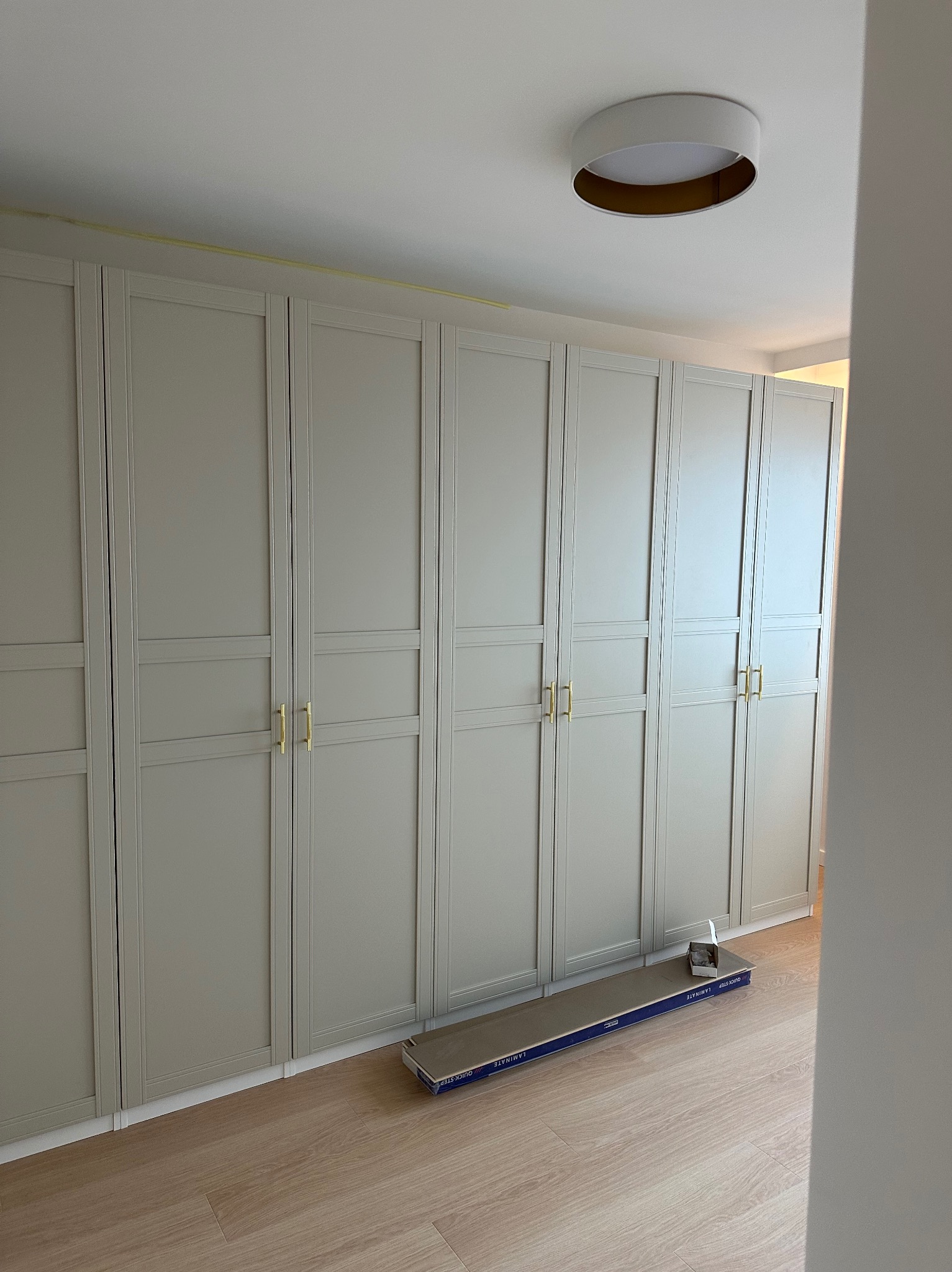 Szary, wbudowany rząd szaf z złotymi uchwytami, maskowany żółtą taśmą sufit, panele podłogowe w opakowaniu na podłodze.