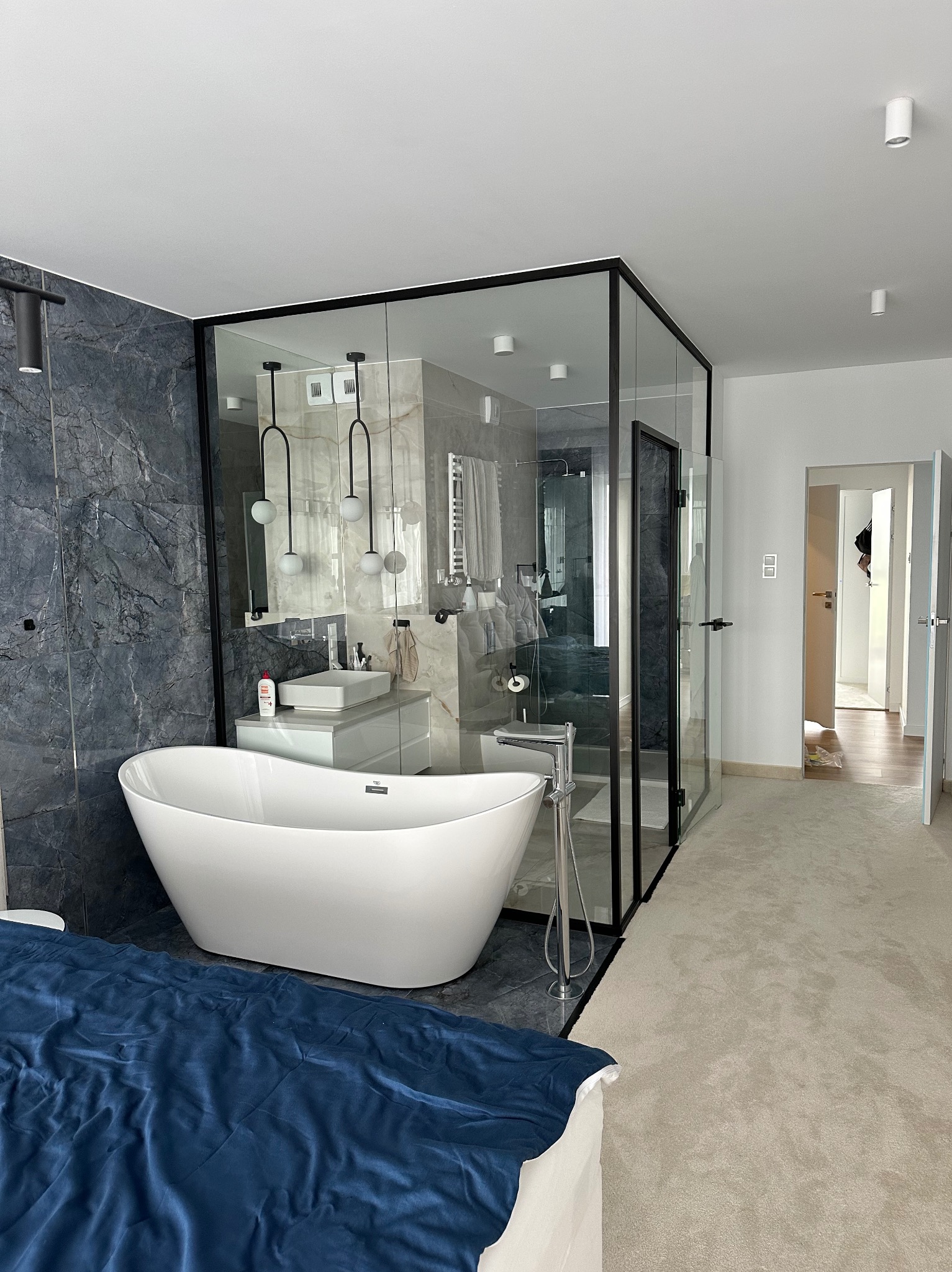 Nowoczesna łazienka z wolnostojącą białą wanną, kabiną prysznicową ze szklanymi ścianami w czarnej ramie, umywalką na szafce i szarą ścianą z marmurowym wzorem, widok z sypialni na wykładzinę...