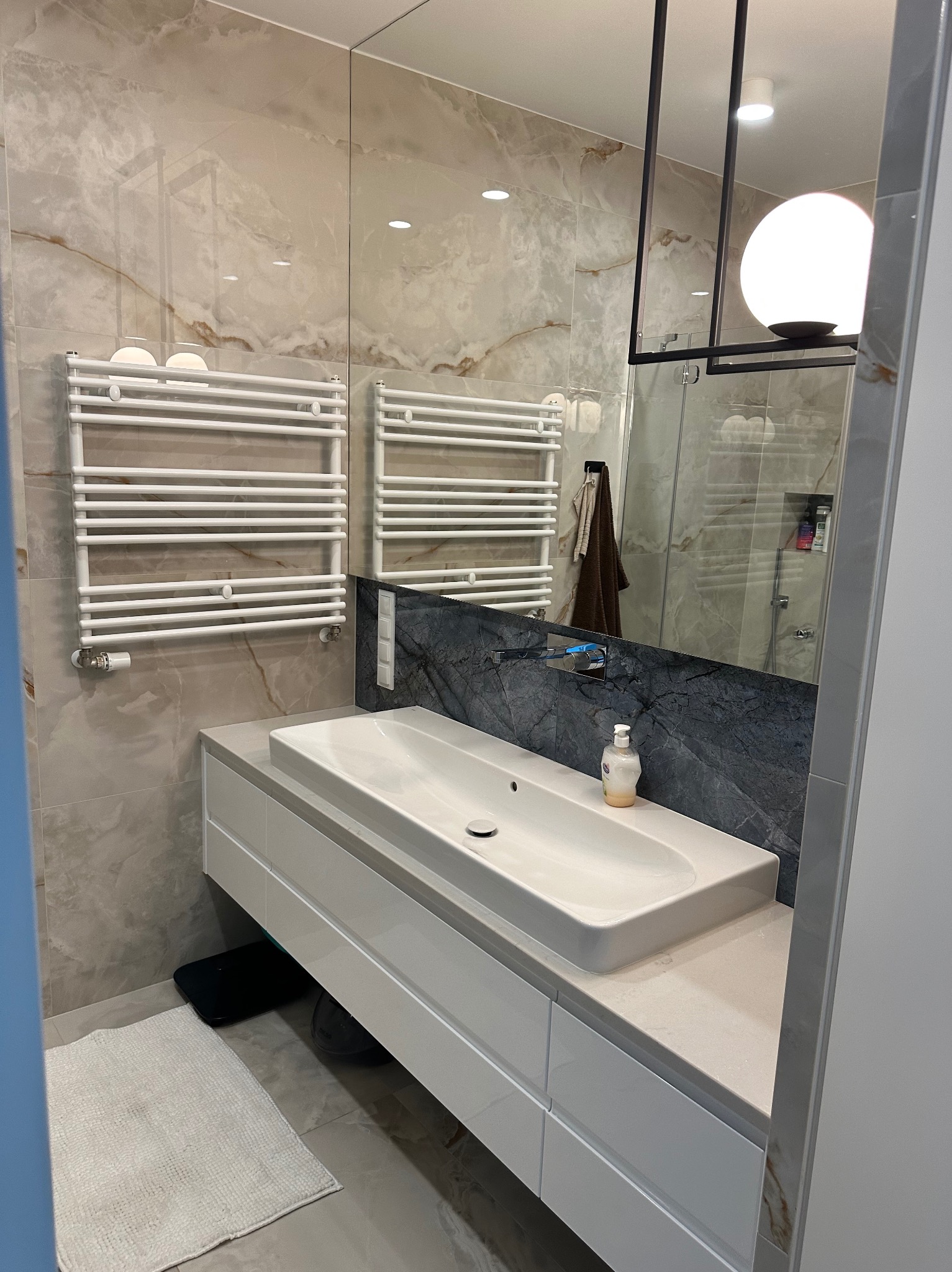 Nowoczesna łazienka w Gdańsku z marmurowymi płytkami, białym blatem umywalkowym i grzejnikiem drabinkowym. W lustrze odbija się kabina prysznicowa.