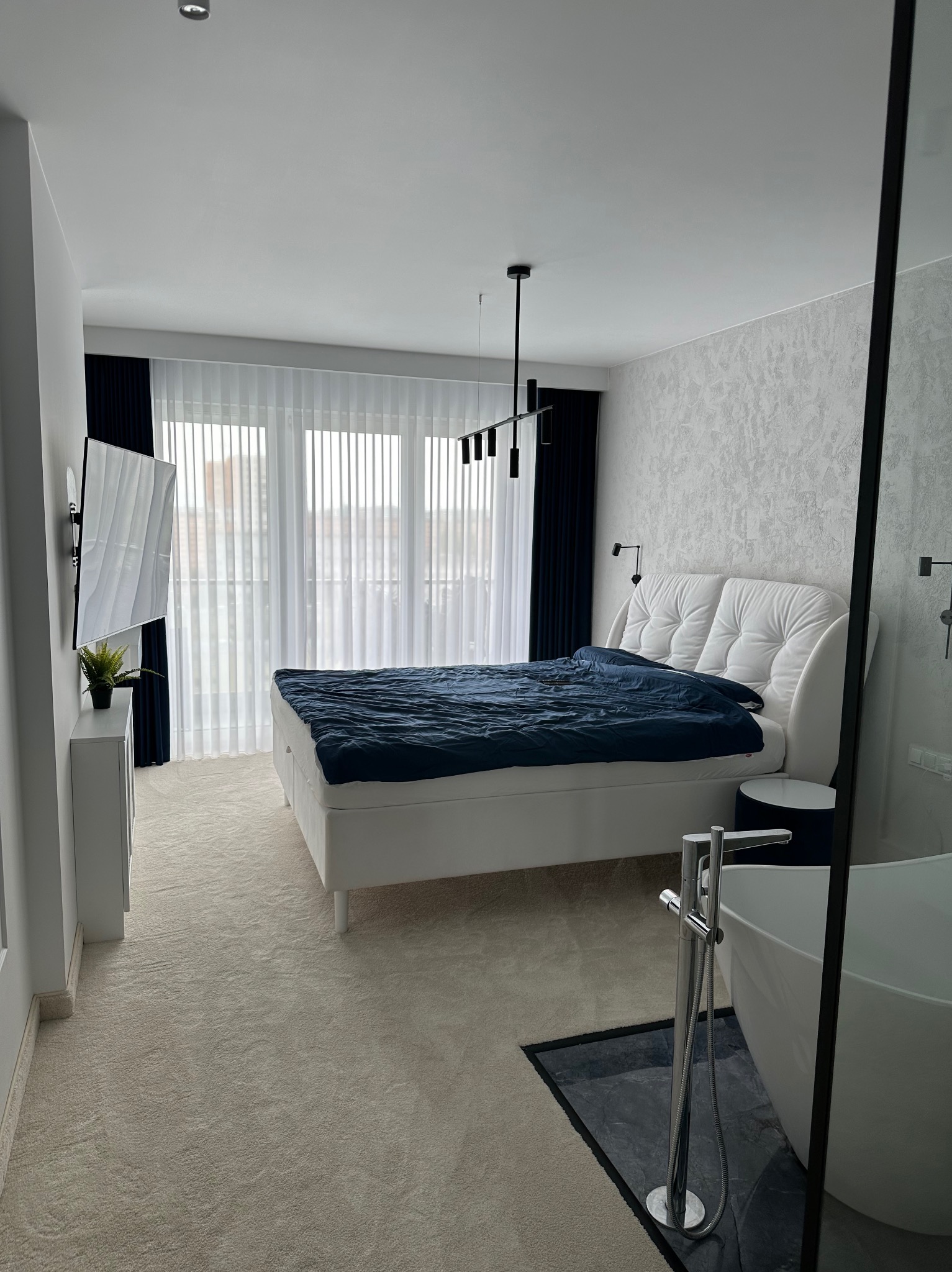 Nowoczesna sypialnia z łazienką: białe łóżko z pikowanym zagłówkiem, granatowa pościel, wisząca lampa, telewizor na ścianie, białe zasłony i wolnostojąca wanna z chromowaną baterią.