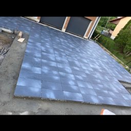 Rubik Eryk Kuźniar - Układanie szarej kostki brukowej na podjeździe do garażu dwustanowiskowego, widoczne niedokończone obrzeża i przygotowane elementy do dalszej pracy.