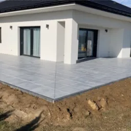 Nowoczesny taras z szarych płytek ceramicznych przy białym domu z czarnym dachem, obrzeża wykończone czarnym kamieniem, widoczne przygotowane podłoże z ziemi i trawy.