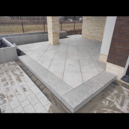 Rubik Eryk Kuźniar - Wyłożony jasnoszarymi płytami taras z obrzeżem z tego samego materiału, widoczne fragmenty elewacji budynku i trawnik w tle.