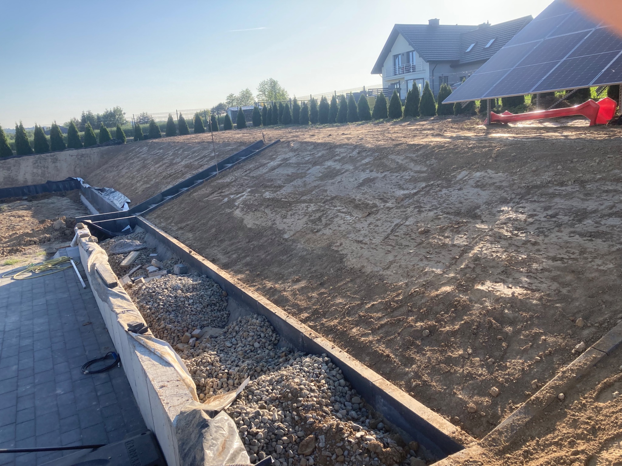 Przygotowany teren pod budowę z wykopem, widoczne obrzeża, częściowo wypełnione drobnym kamieniem, w tle dom jednorodzinny i panele solarne.
