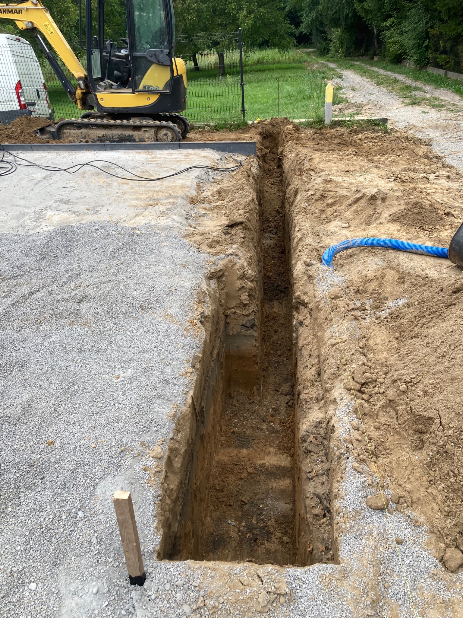 Wykopany wąski rów w ziemi, częściowo zasypany żwirem, z widoczną niebieską rurą karbowaną i żółtą mini koparką Yanmar w tle, teren przygotowany pod instalacje.