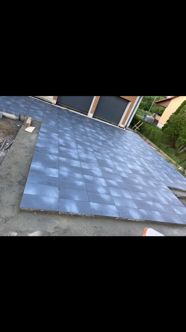 Układanie szarej kostki brukowej na podjeździe do garażu dwustanowiskowego, widoczne niedokończone obrzeża i przygotowane elementy do dalszej pracy.