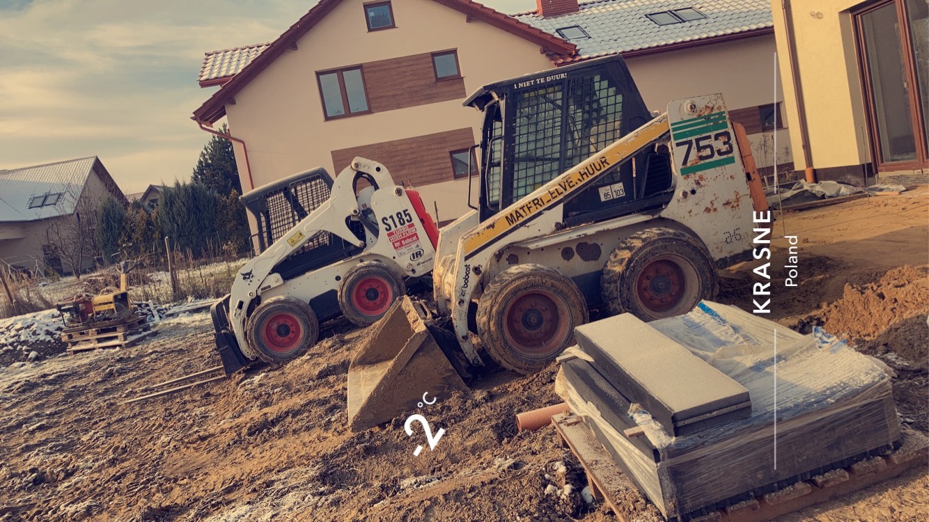 Dwa miniładowarki Bobcat na placu budowy w słoneczny, mroźny dzień, w tle dom w stanie surowym zamkniętym i ubijarka na palecie, obok stos kostki brukowej w folii.