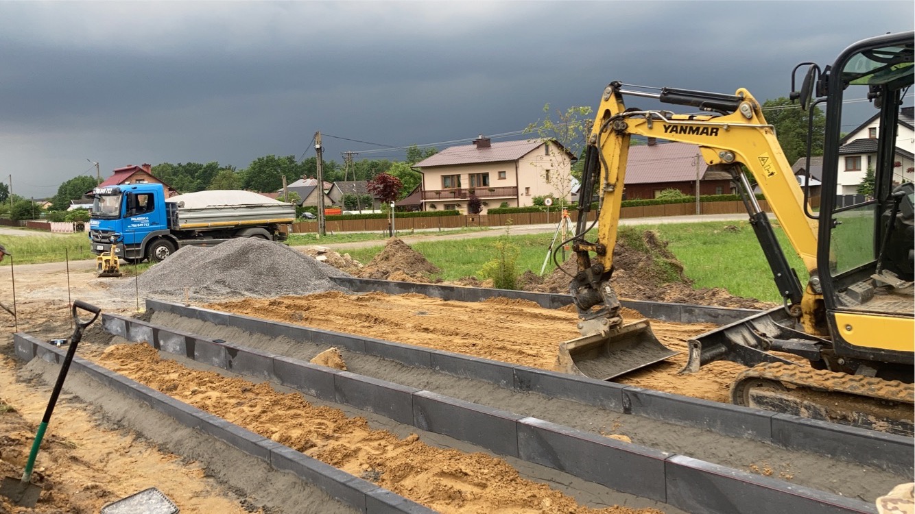 Żółta mini koparka Yanmar w trakcie przygotowywania podłoża pod nawierzchnię z kostki brukowej, widoczny wykop, krawężniki, piasek i samochód ciężarowy z żwirem w tle.