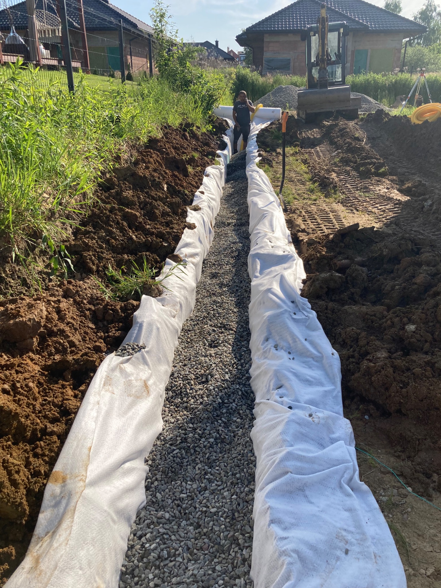 Wykop w ziemi wyłożony białą geowłókniną, wypełniony drobnym kruszywem, w tle koparka i pracownik w trakcie robót ziemnych.