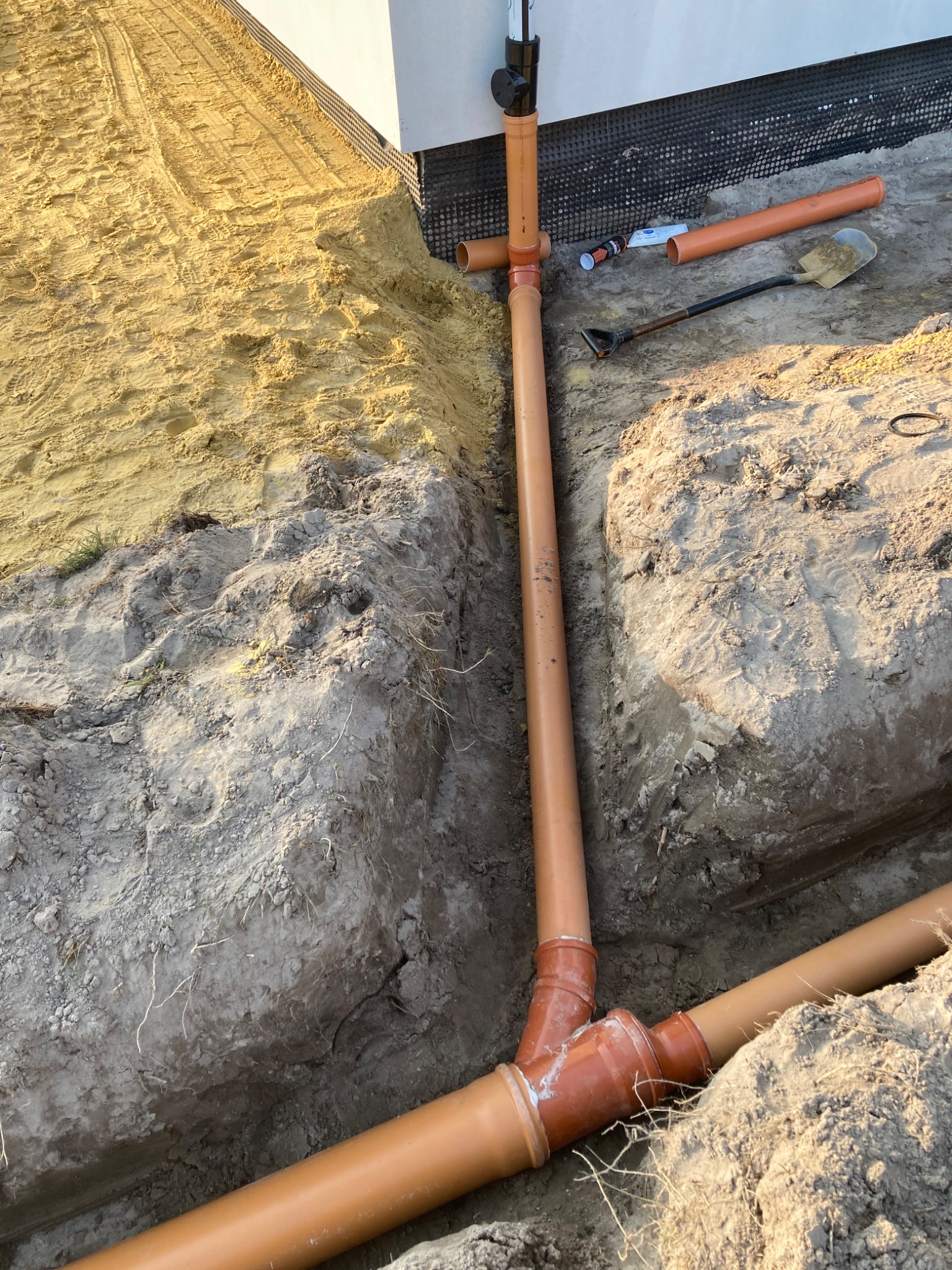 Pomarańczowe rury kanalizacyjne ułożone w wykopie ziemnym, widoczna łopata i inne narzędzia, piasek oraz zabezpieczenie budynku.