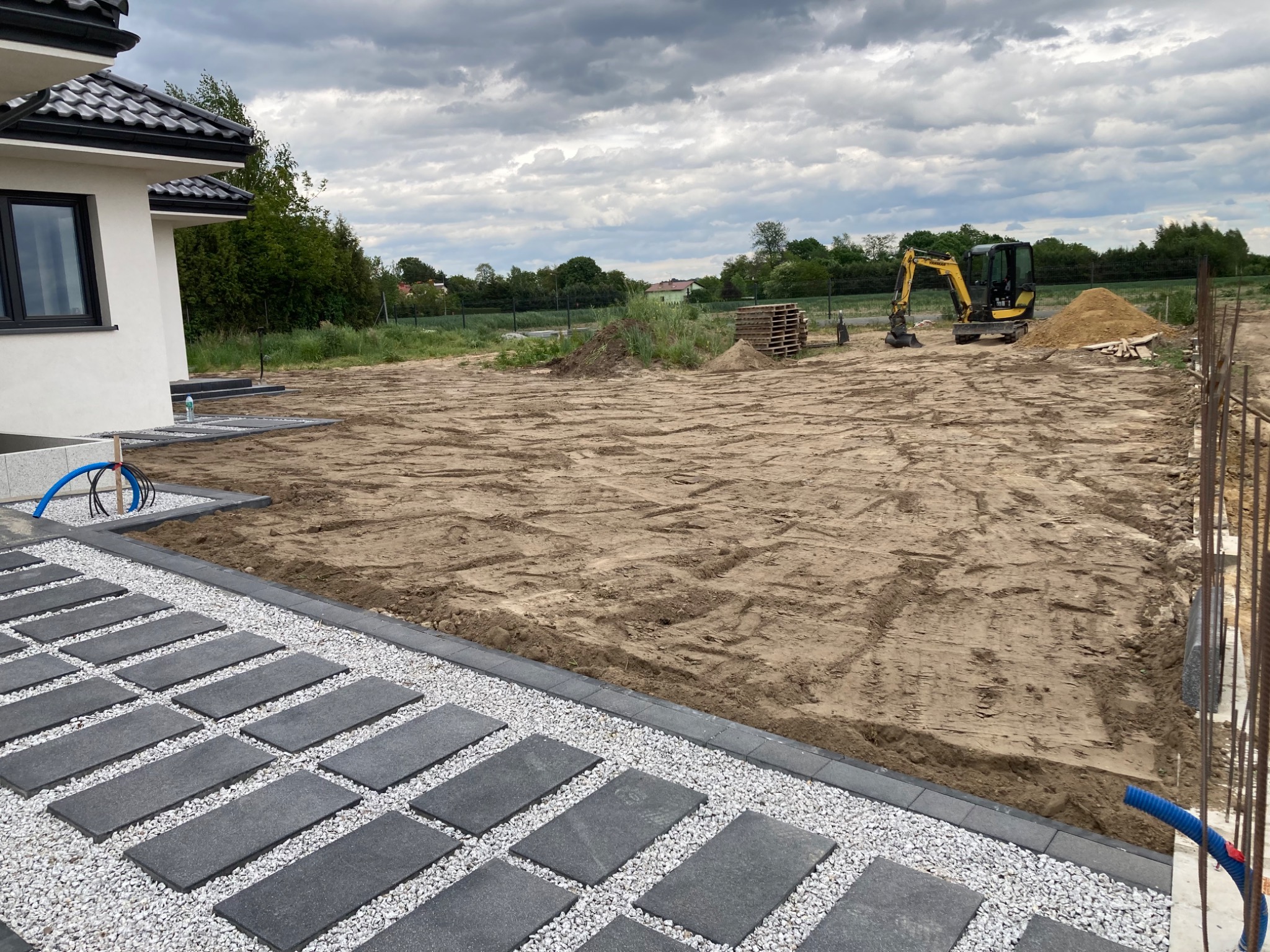 Przygotowany teren pod budowę domu z ułożonym chodnikiem z płyt i podsypką z białego żwiru, widoczny minikoparka, palety i sterta ziemi pod pochmurnym niebem.