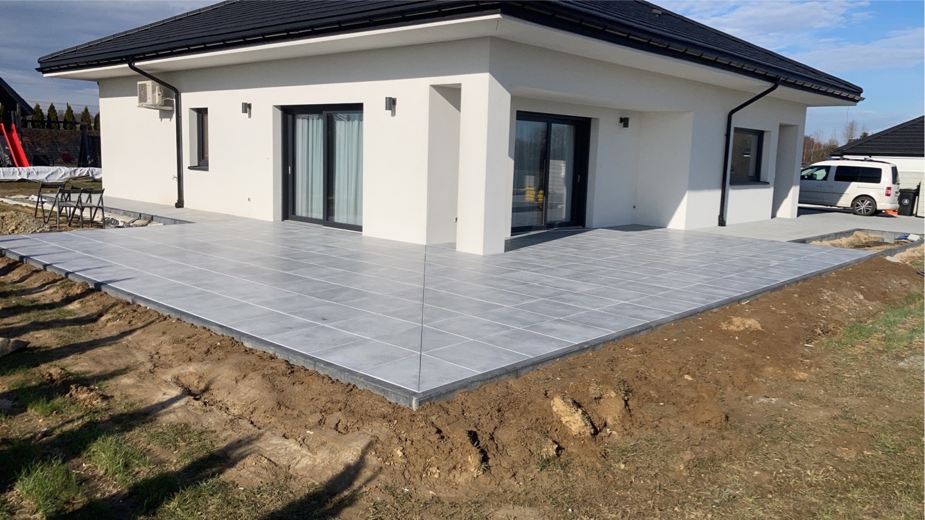 Nowoczesny taras z szarych płytek ceramicznych przy białym domu z czarnym dachem, obrzeża wykończone czarnym kamieniem, widoczne przygotowane podłoże z ziemi i trawy.