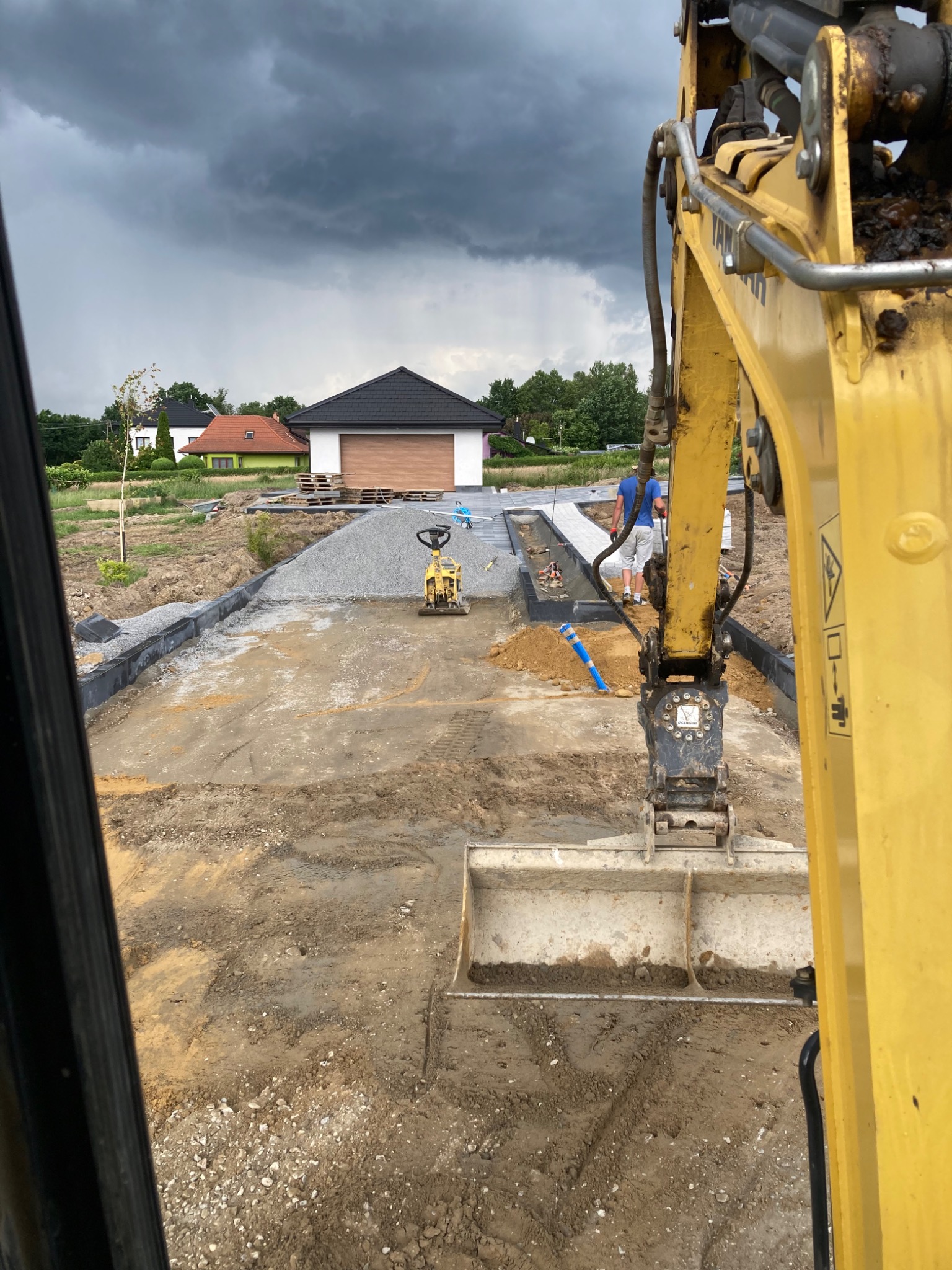 Widok z kabiny koparki na przygotowywany teren pod budowę drogi dojazdowej, widoczny ubijak wibracyjny, krawężniki i ułożona kostka brukowa, w tle domy pod pochmurnym niebem.