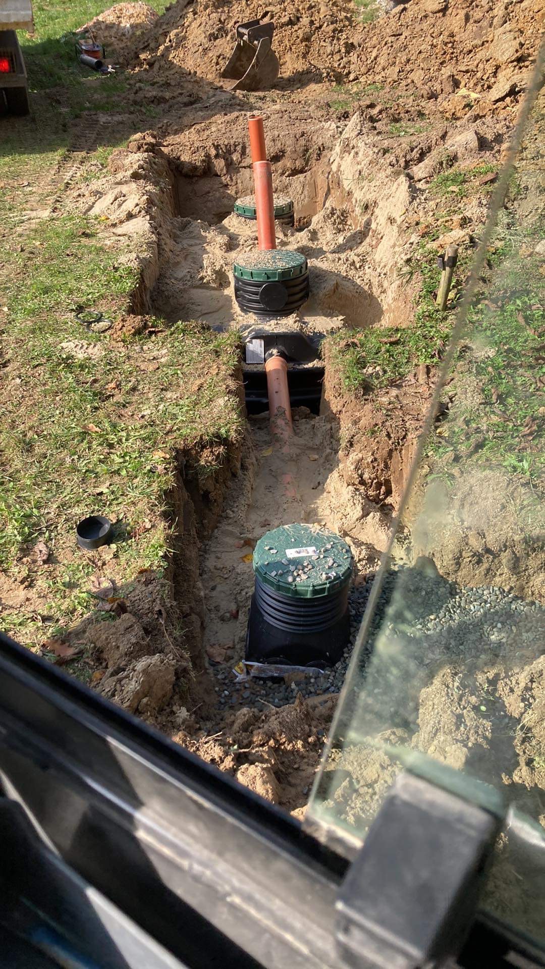 Wykop z ułożonymi rurami i studzienkami kanalizacyjnymi z zielonymi pokrywami, widok z kabiny maszyny budowlanej.