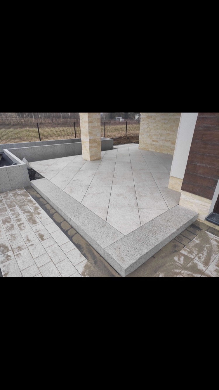 Wyłożony jasnoszarymi płytami taras z obrzeżem z tego samego materiału, widoczne fragmenty elewacji budynku i trawnik w tle.