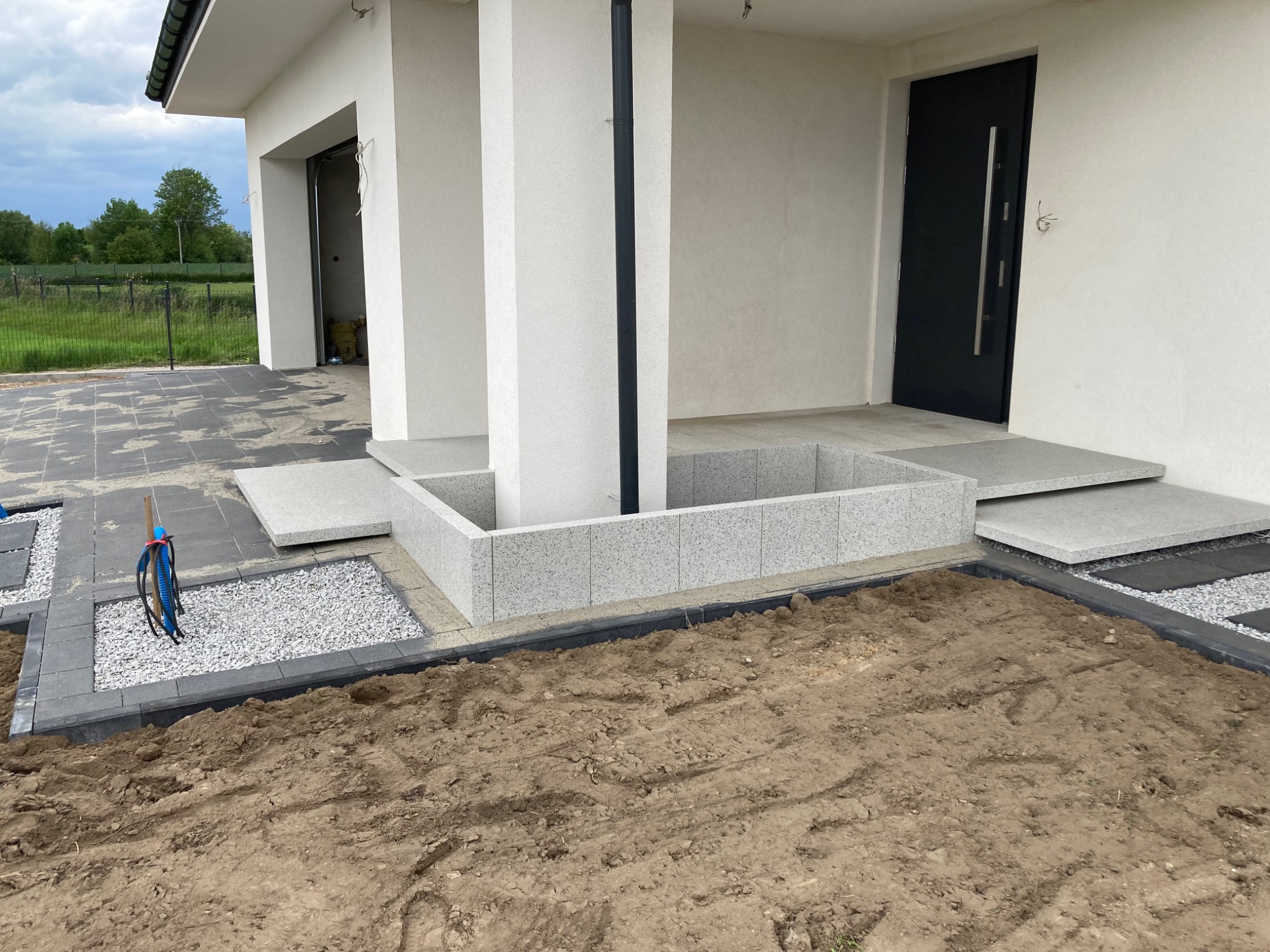 Układanie kostki brukowej i płyt chodnikowych wokół domu z elementami drenażu, widoczne przygotowane podłoże z ziemi i kabel w niebieskiej osłonie.