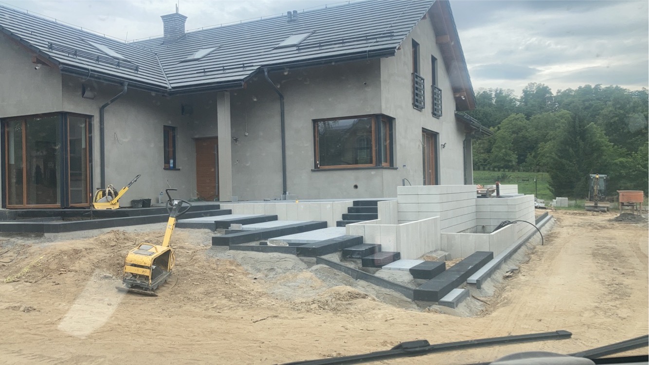Budowa domu jednorodzinnego w trakcie wykańczania terenu: wyrównywanie gruntu zagęszczarkami, układanie schodów i obrzeży z kostki brukowej, widoczne fundamenty tarasu z bloczków betonowych.