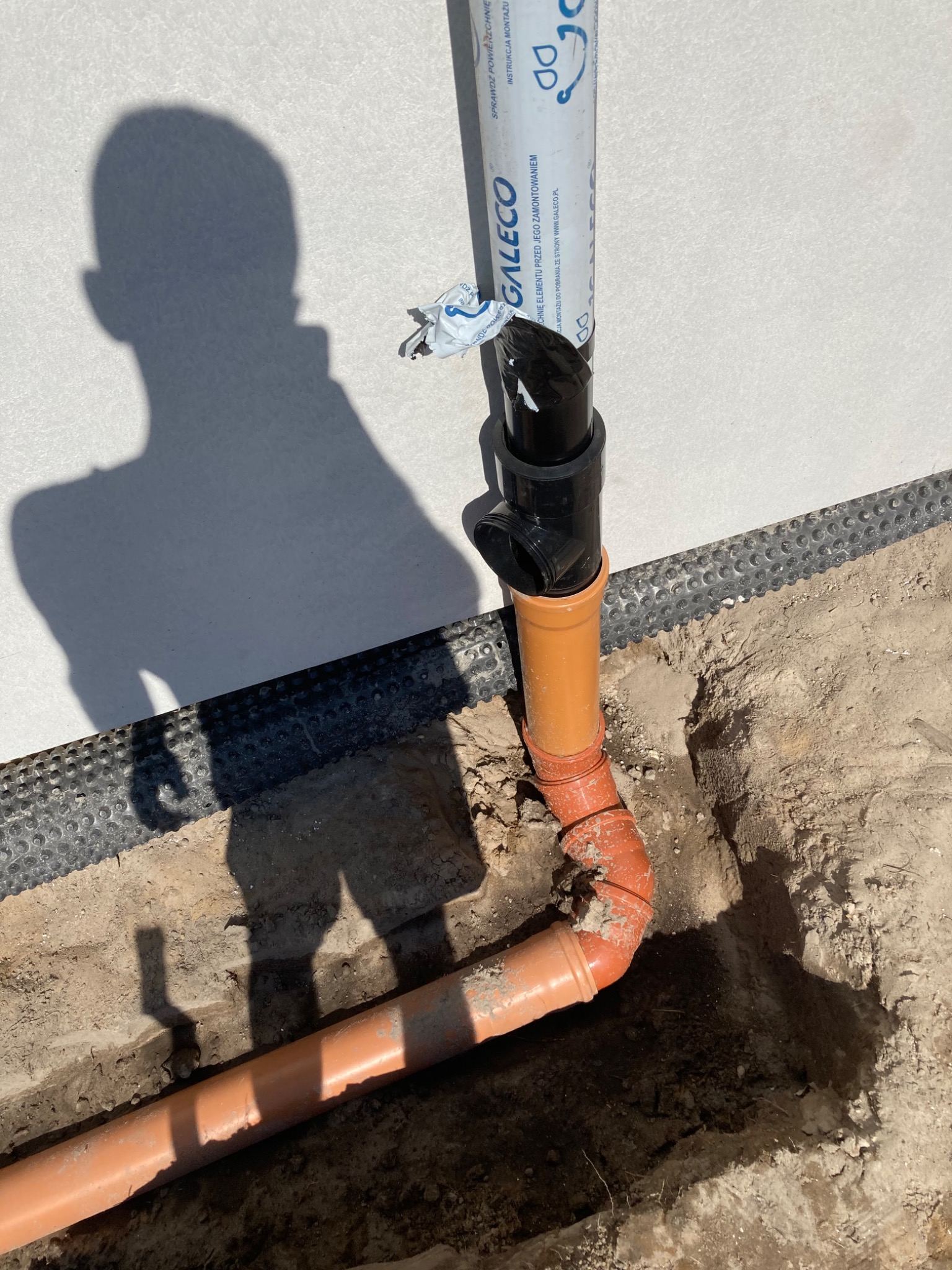 Rury kanalizacyjne z tworzywa sztucznego, pomarańczowe i czarne, wystające z wykopu w ziemi, częściowo zakopane, z widocznym cieniem osoby fotografującej na ścianie budynku w tle.
