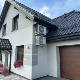 Dwa białe klimatyzatory zamontowane na elewacji domu jednorodzinnego z brązowym dachem i garażem, widoczne kwiaty w oknach.