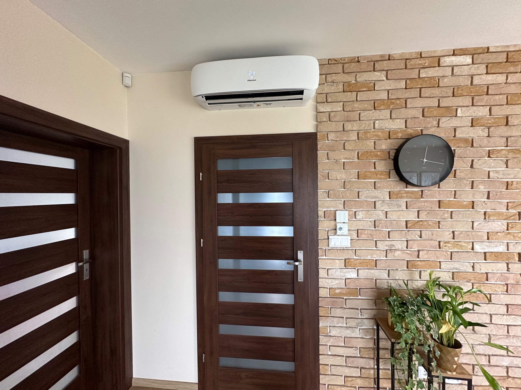 Biały klimatyzator Hisense zamontowany nad ciemnobrązowymi drzwiami z poziomymi przeszkleniami, na ścianie z cegły dekoracyjnej, obok zegar i rośliny doniczkowe.