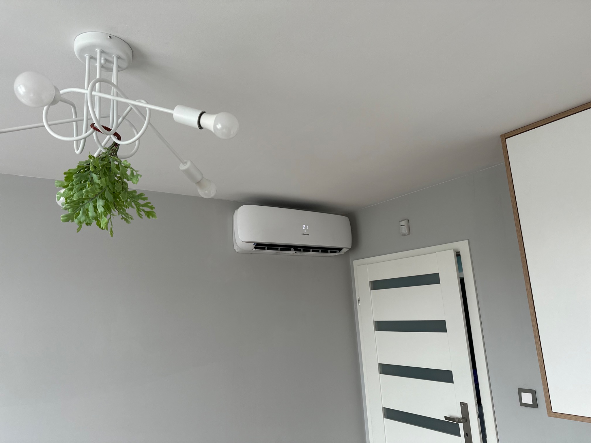 Biały klimatyzator Hisense zamontowany na szarej ścianie w pokoju, pod nowoczesną lampą z zielonymi roślinami