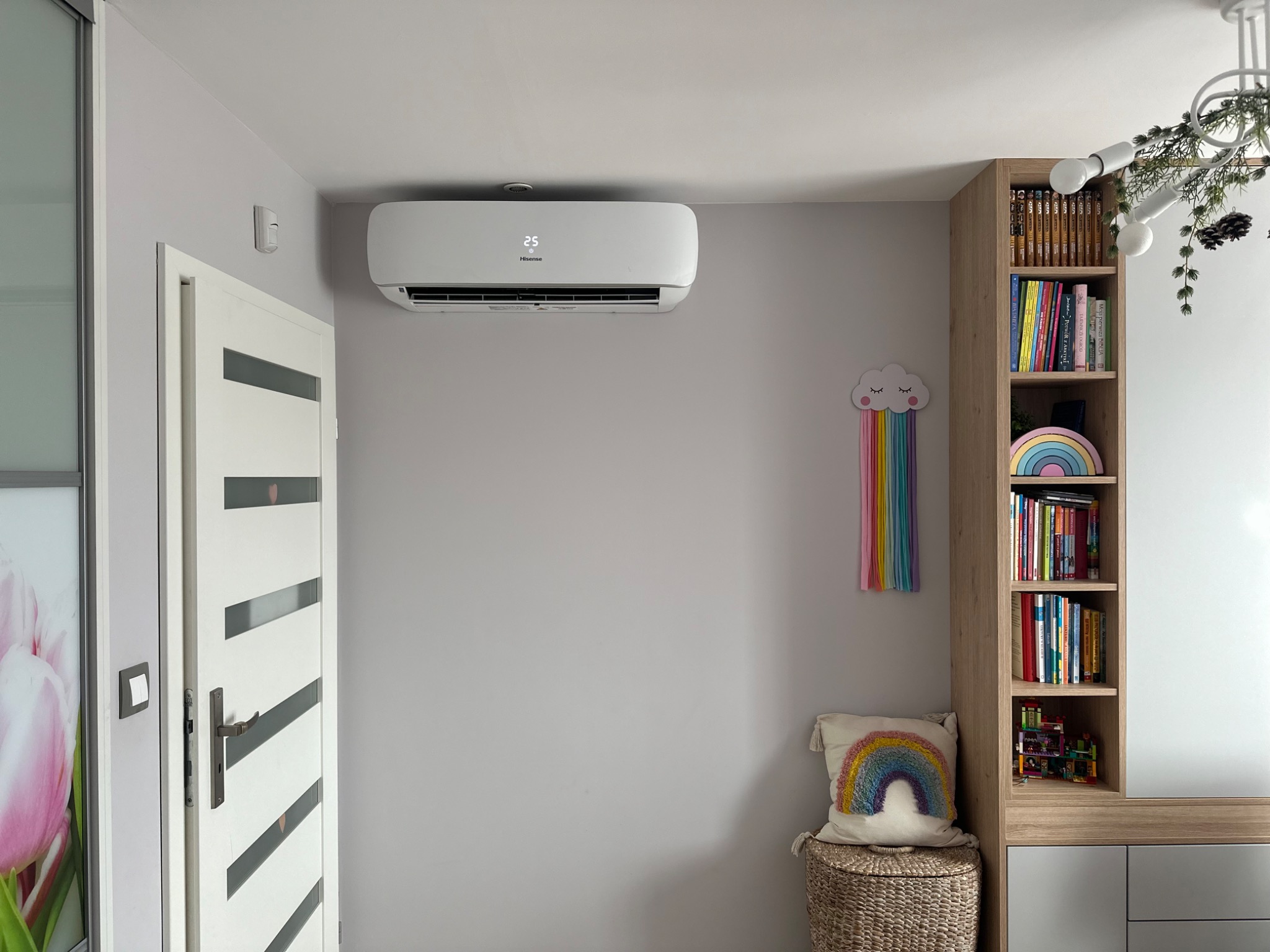 Biały klimatyzator Hisense zamontowany na szarej ścianie w pokoju dziecięcym, widoczny wyświetlacz z temperaturą 25 stopni, obok regał z książkami i dekoracje w kształcie tęczy.