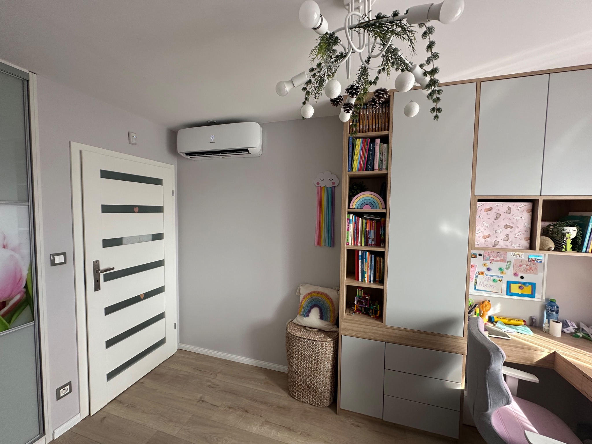 Biały klimatyzator ścienny w pokoju dziecięcym z regałem na książki i biurkiem, widok z niskiego kąta.